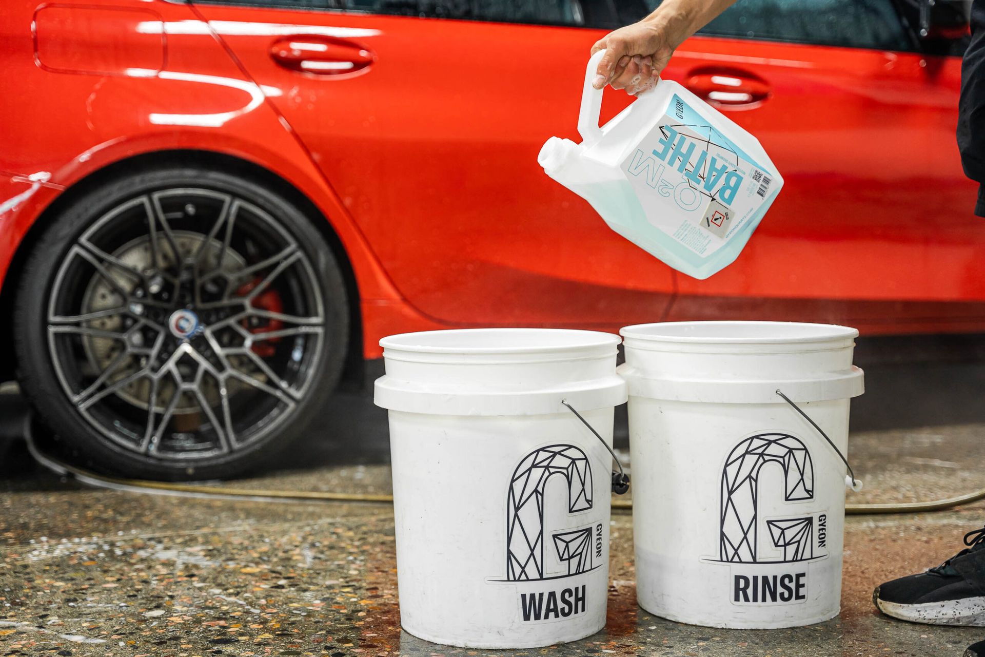 A person is pouring liquid into two white buckets next to a red car.