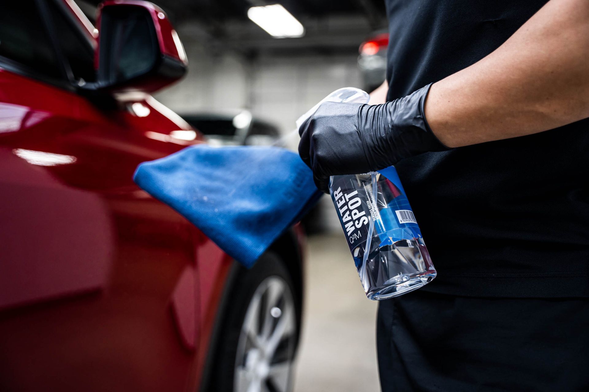 A person is holding a blue towel and a spray bottle in front of a red car.