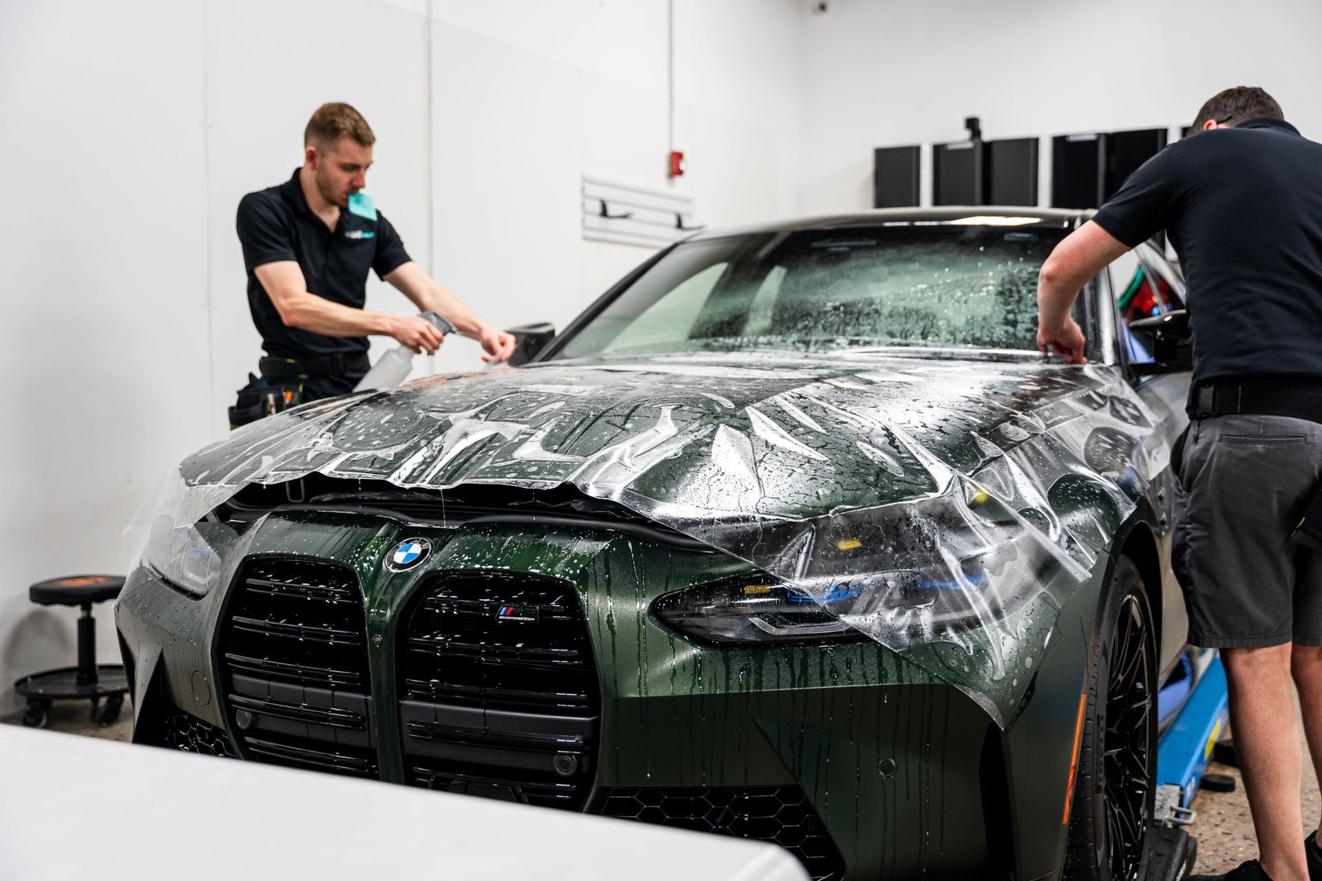 Two men are wrapping a car in plastic in a garage.