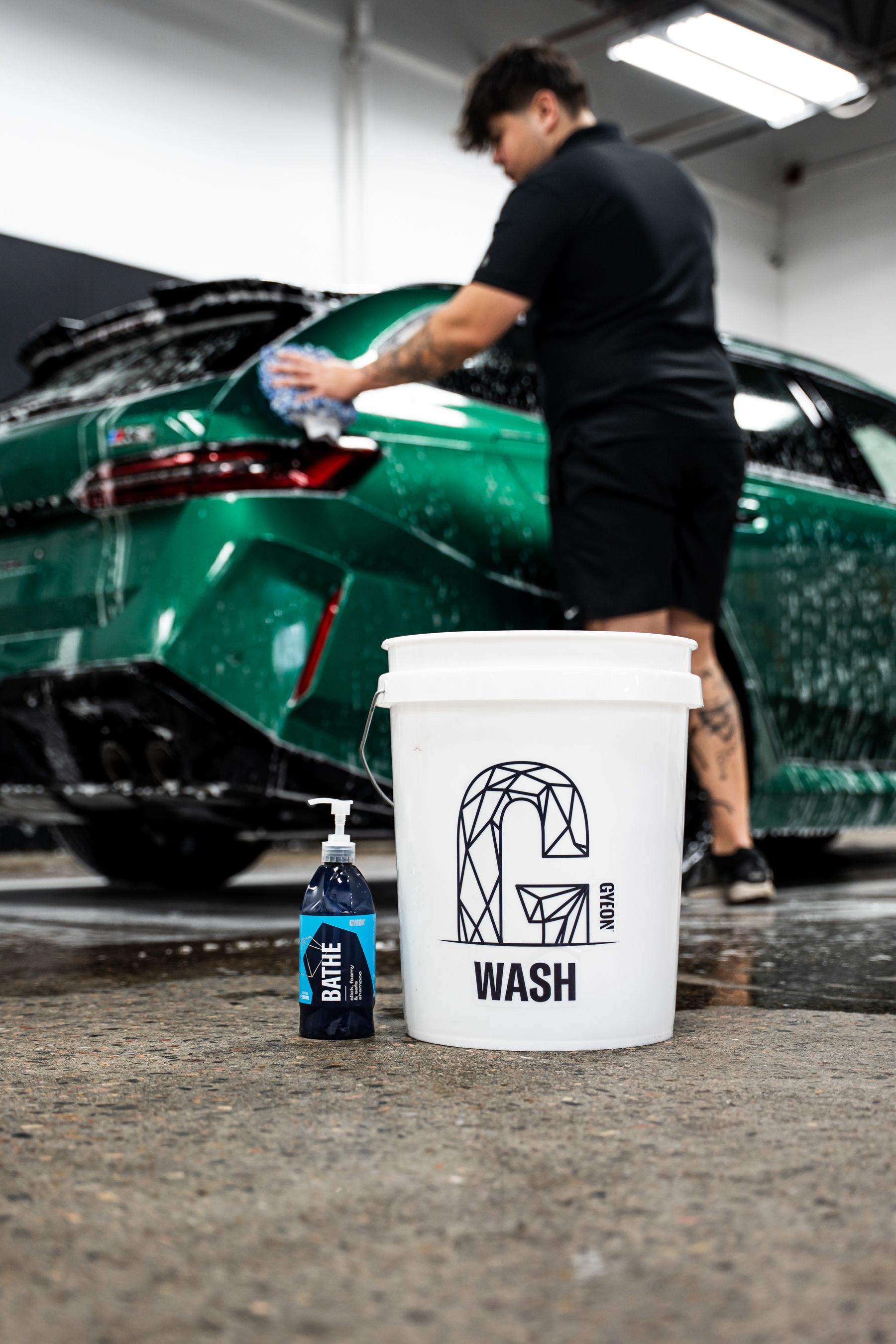 A person washing a green car with soap in a garage; bucket and soap bottle in foreground.