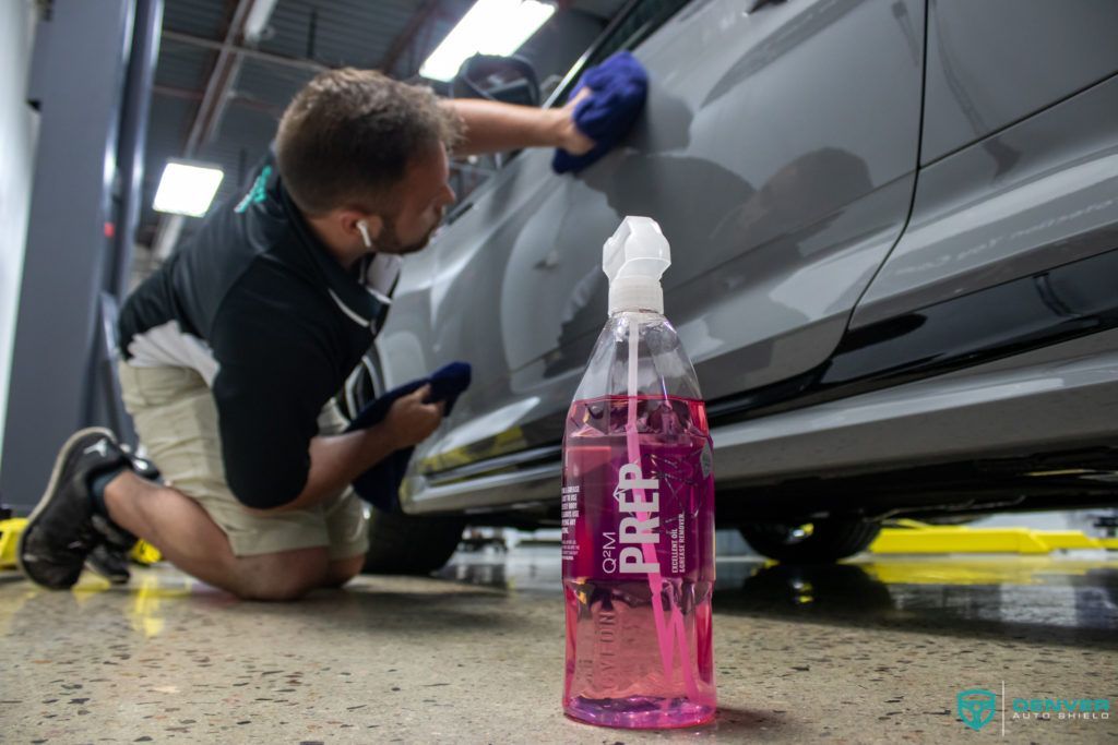 A man is kneeling down to polish a car in a garage next to a bottle of prep.