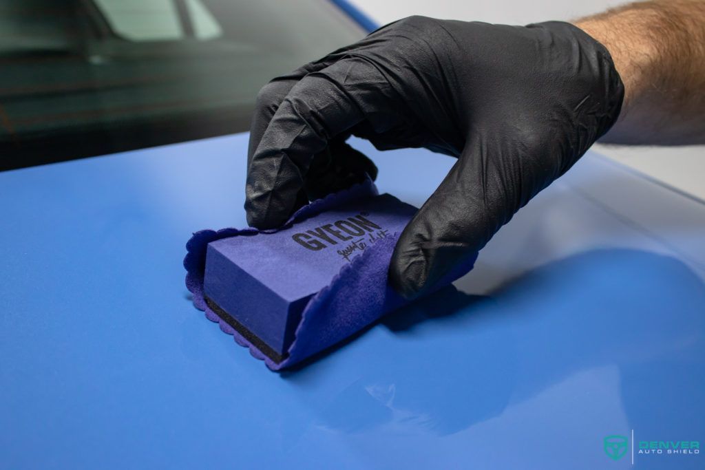 A person wearing black gloves is polishing a blue car with a purple sponge.