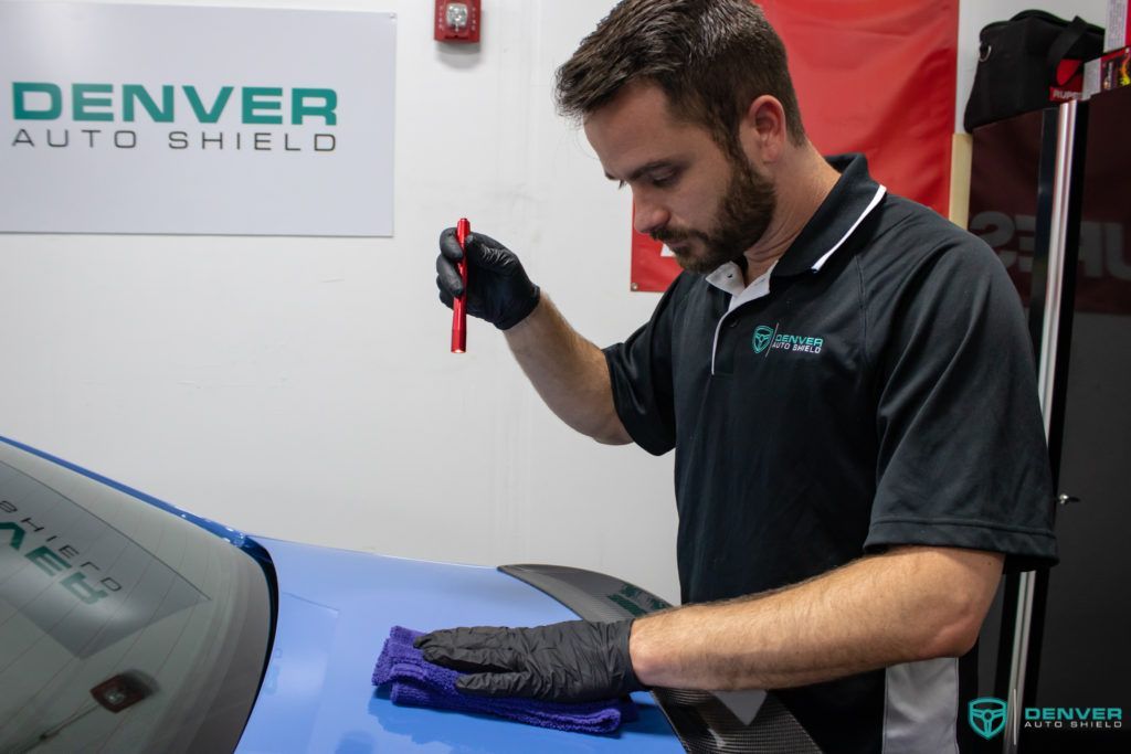 A man is working on a car with a denver auto shield sign in the background.