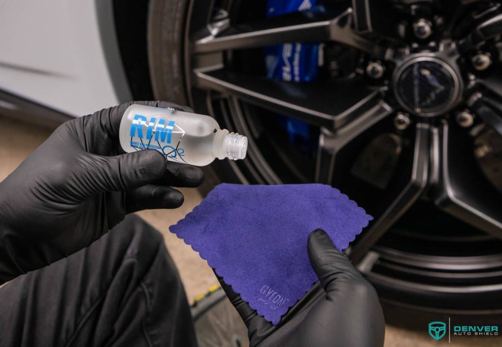 A person is holding a bottle of ceramic coating and a cloth in front of a car wheel.