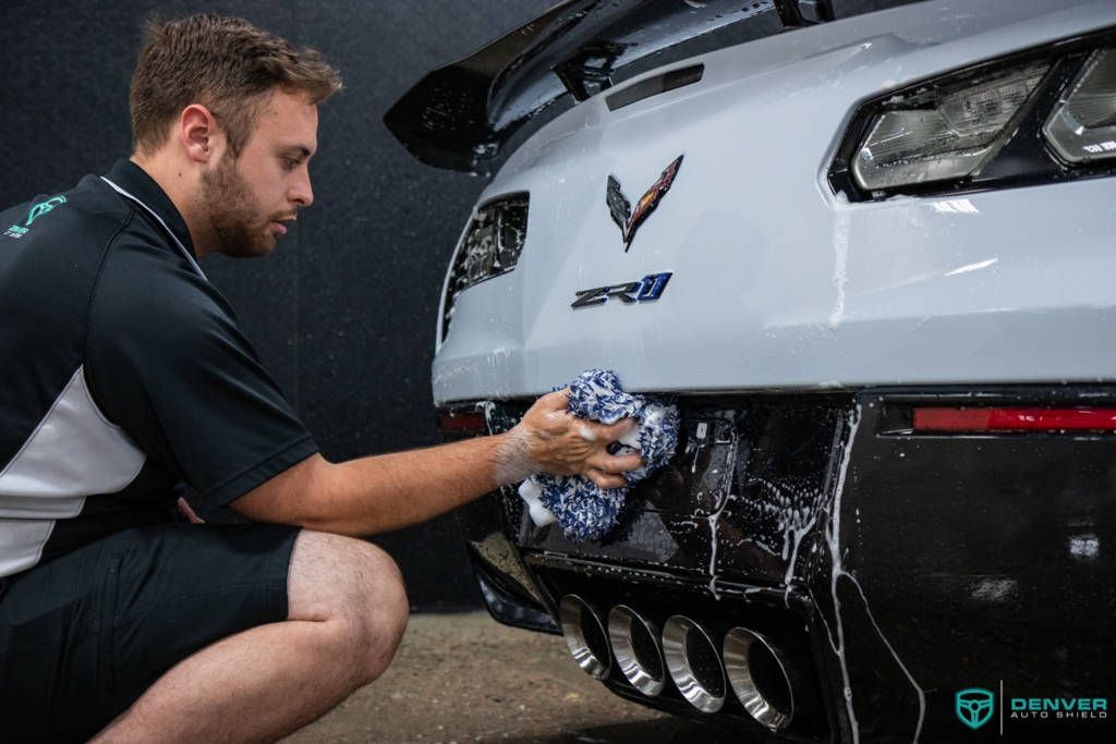 A man is cleaning the back of a white corvette with a cloth.