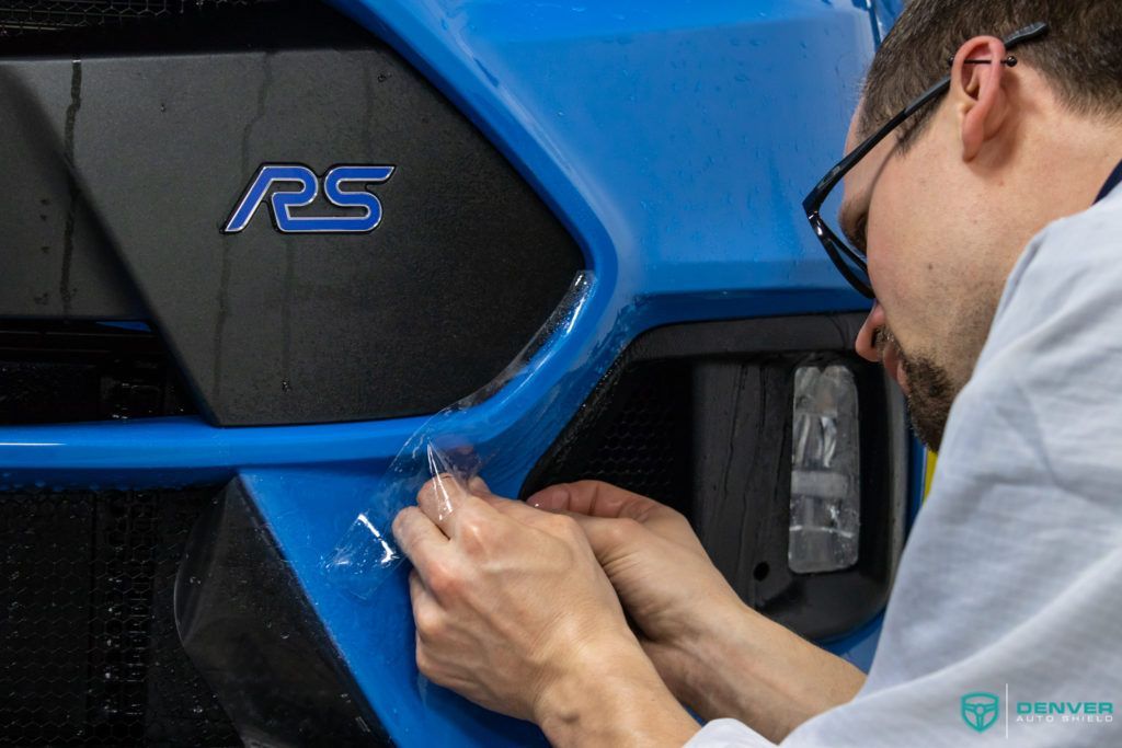 A man is applying a protective film to the front bumper of a blue car.