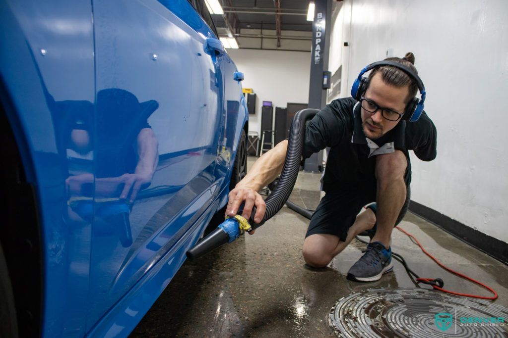 A man wearing headphones is kneeling down next to a blue car.