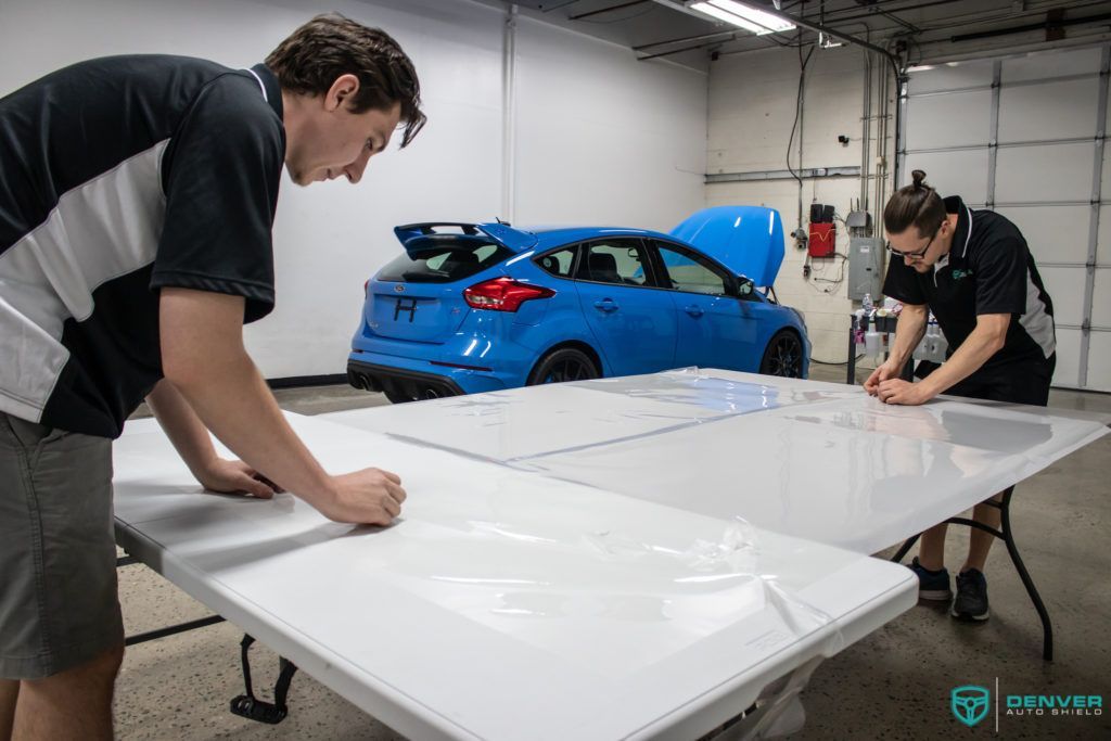 Two men are working on a table with a blue car in the background.