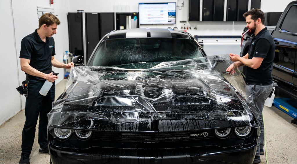 Two men are wrapping a black dodge challenger with clear plastic.