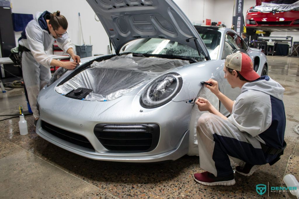 Two men are wrapping a silver sports car in a garage.