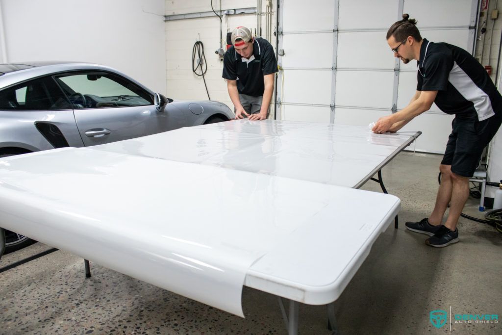 Two men are working on a table in a garage next to a car.