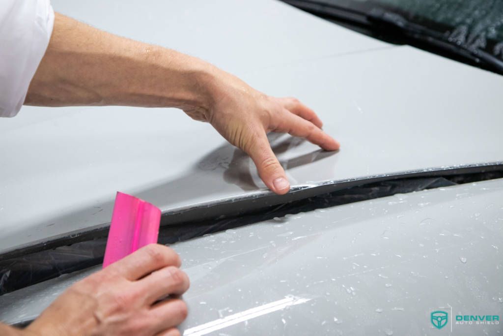 A person is using a pink squeegee to clean the hood of a car.