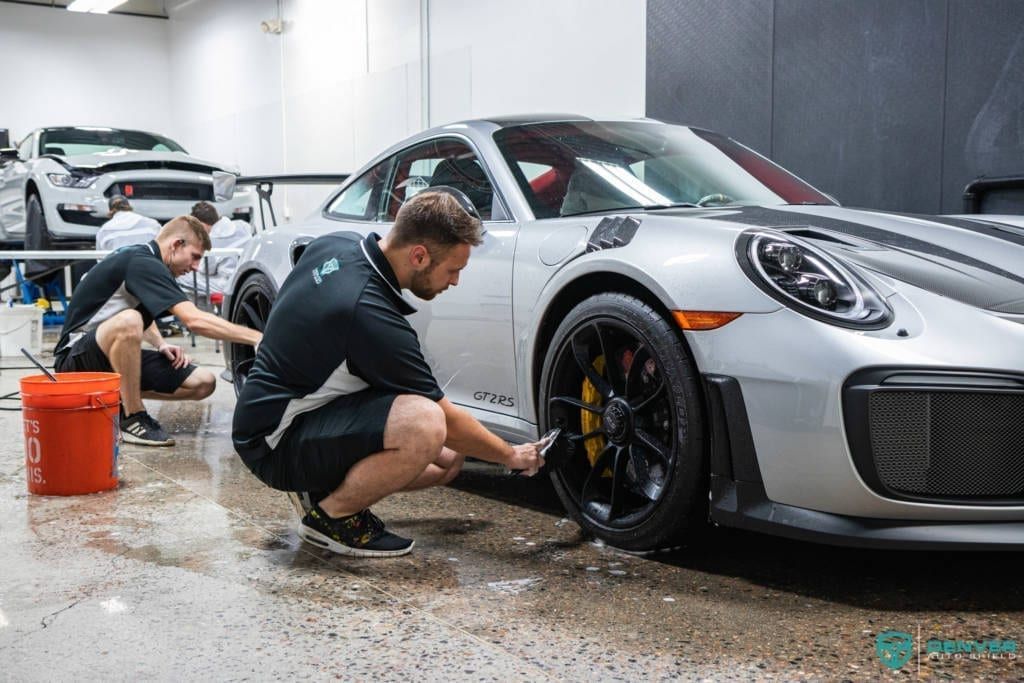 A man is kneeling down next to a silver sports car in a garage.