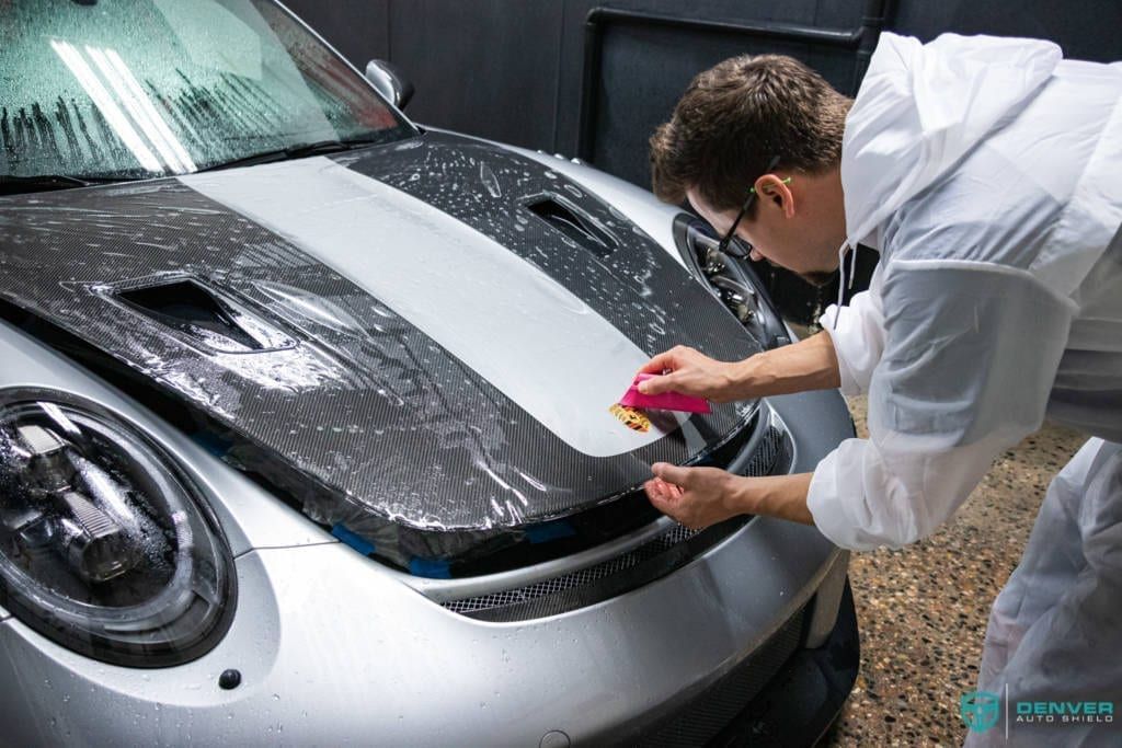 A man is covering the hood of a silver car with a plastic wrap.