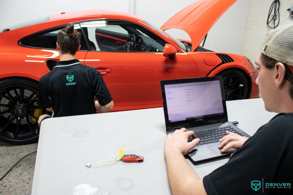 A man is sitting at a table with a laptop in front of a red car.