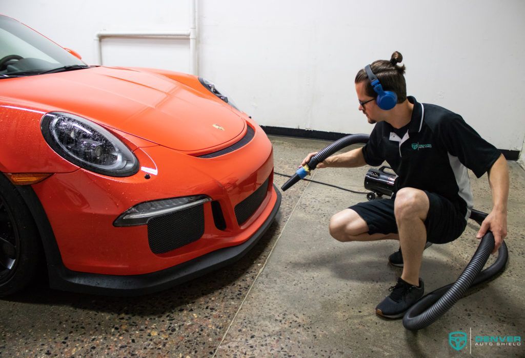 A man is cleaning the front of an orange porsche with a vacuum cleaner.