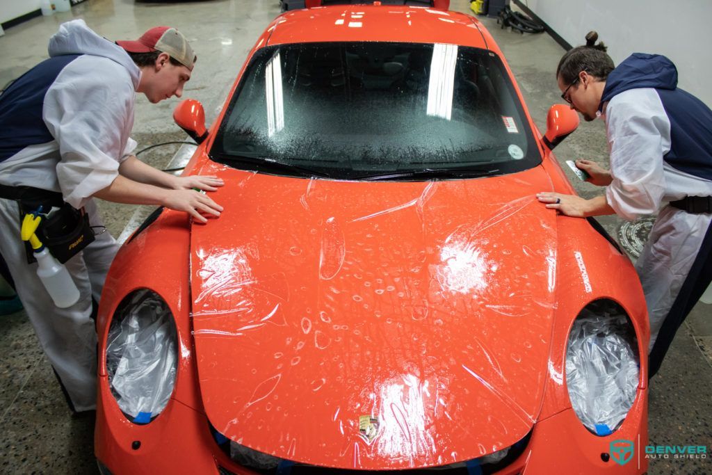 Two men are wrapping an orange sports car with plastic wrap.