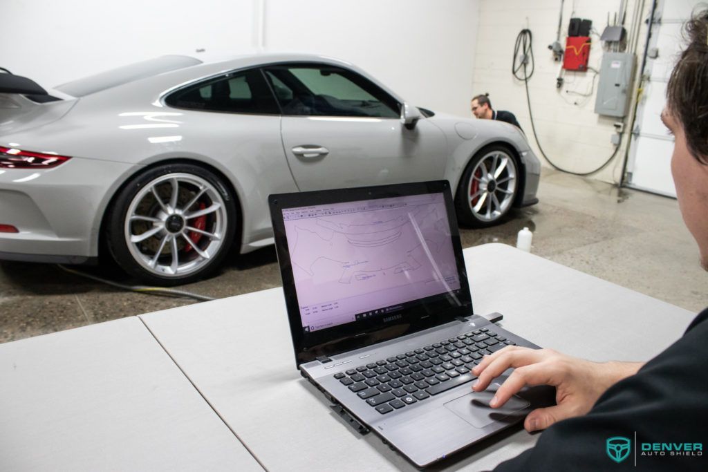 A man is using a laptop computer in front of a silver car.