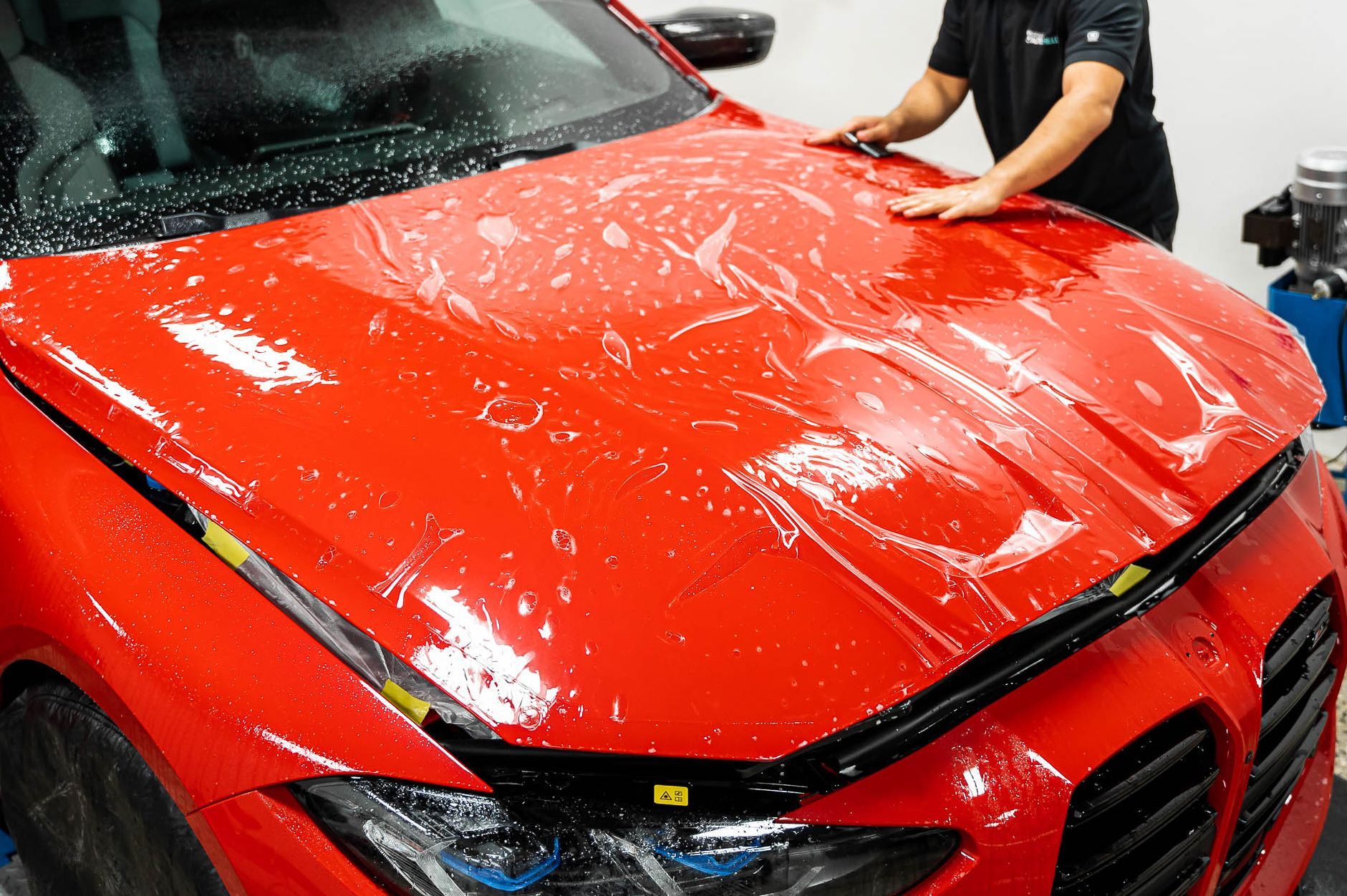 A man is covering the hood of a red car with plastic wrap.