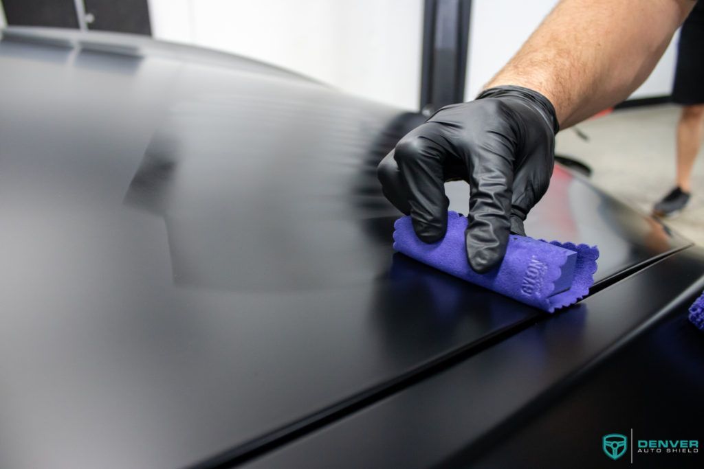 A person wearing black gloves is cleaning a black car with a purple cloth.