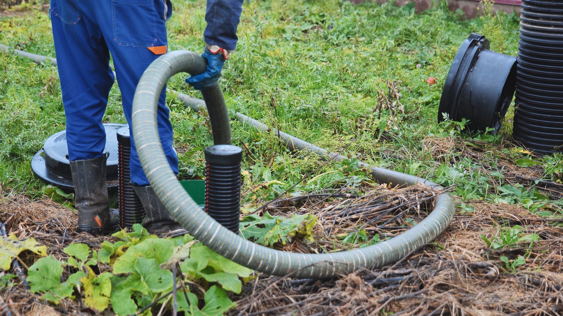 Worker in blue pants uses a hose to clear mud from a grassy trench outdoors. Worker in blue pants uses a hose to clear mud from a grassy trench outdoors.