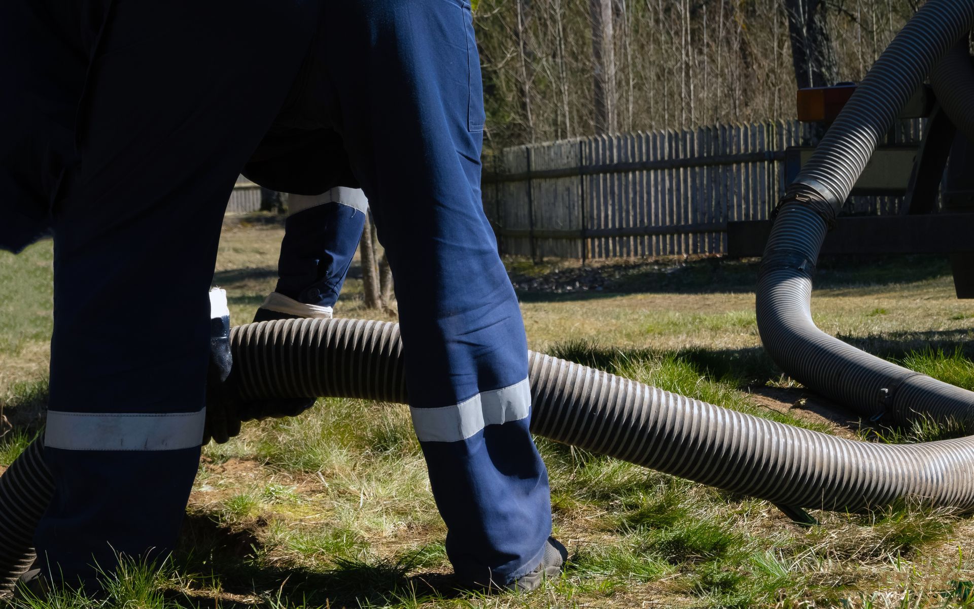 Workers in blue pants handling large black hoses on grass near a fence