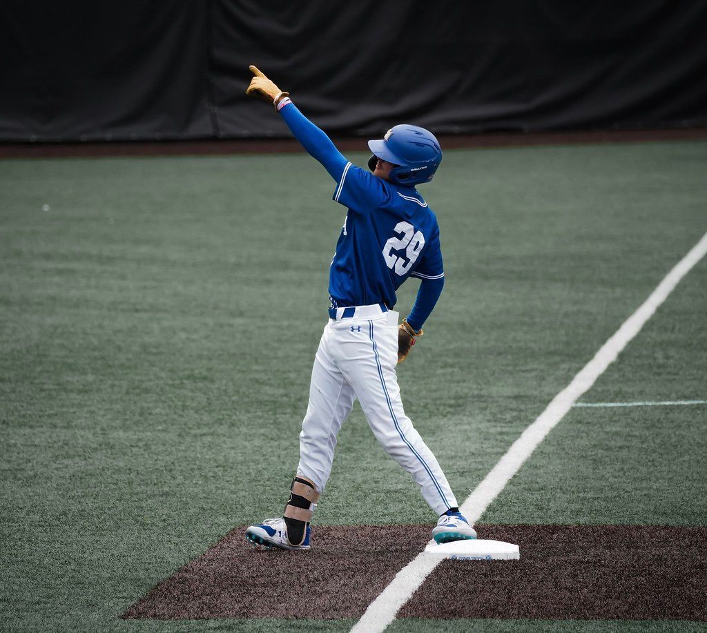 Baseball player in blue uniform points upward on a field near first base.