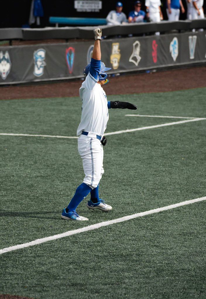 Baseball player in white and blue uniform raises glove on field near the dugout