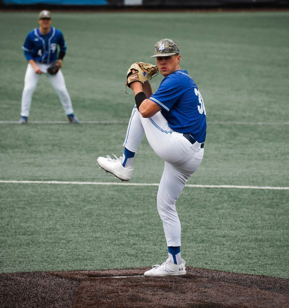 Baseball pitcher in blue uniform mid-windup on the mound, with an infielder in the background.