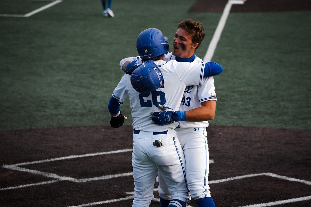 Two baseball players in white and blue uniforms hug on the field after a play.