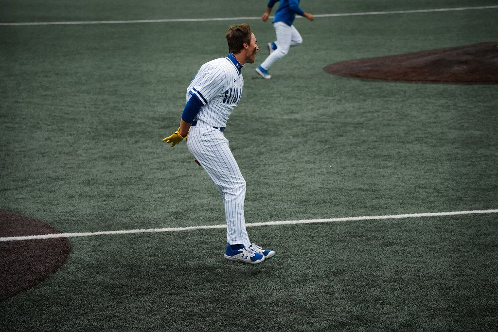Baseball player in white pinstripes standing on a field, with another player running in the background.