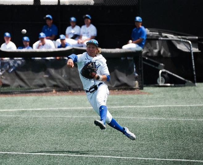 Baseball player in white and blue sprinting on the field, with teammates seated in the dugout behind.