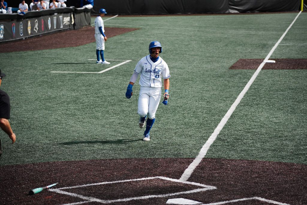 Baseball player in white uniform walking near home plate on a turf field, with another player in the background