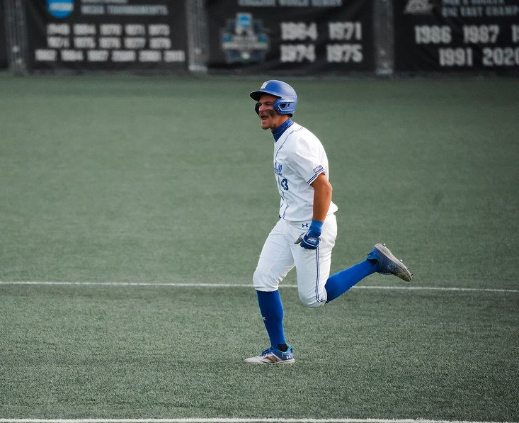 Baseball player in white and blue uniform running on a turf field, with outfield scoreboard in the background