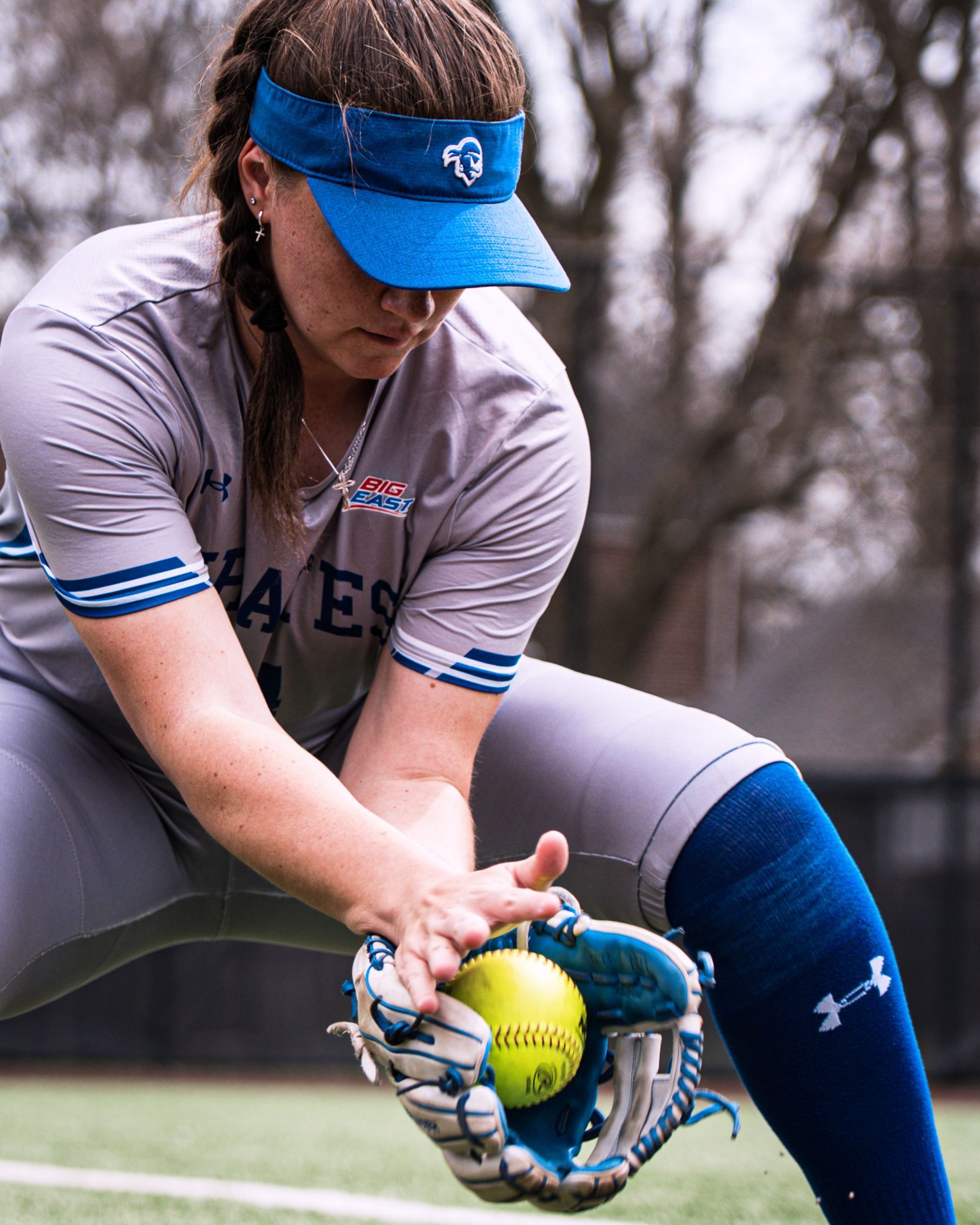 Softball player in gray uniform and blue visor fielding a ball in a glove on the field