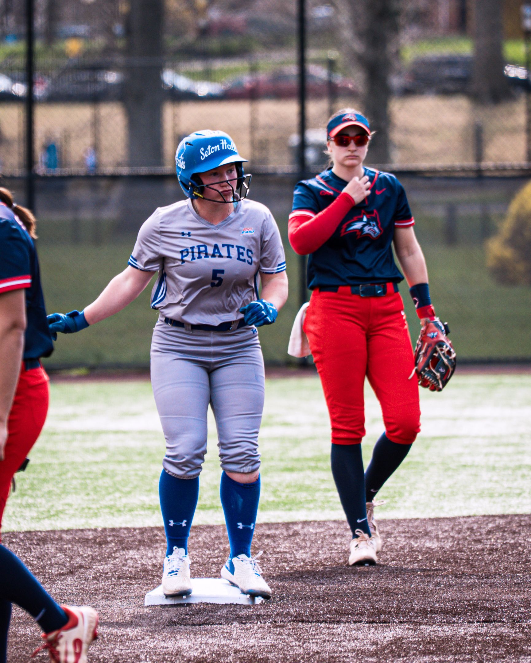 Two softball players on a field, one in gray uniform and blue helmet, one in red uniform behind her.