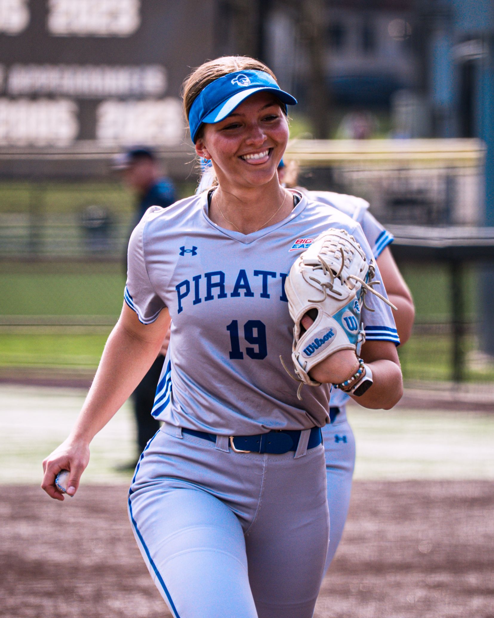 Softball player in light blue Pirates uniform smiling on the field with a glove on her left hand