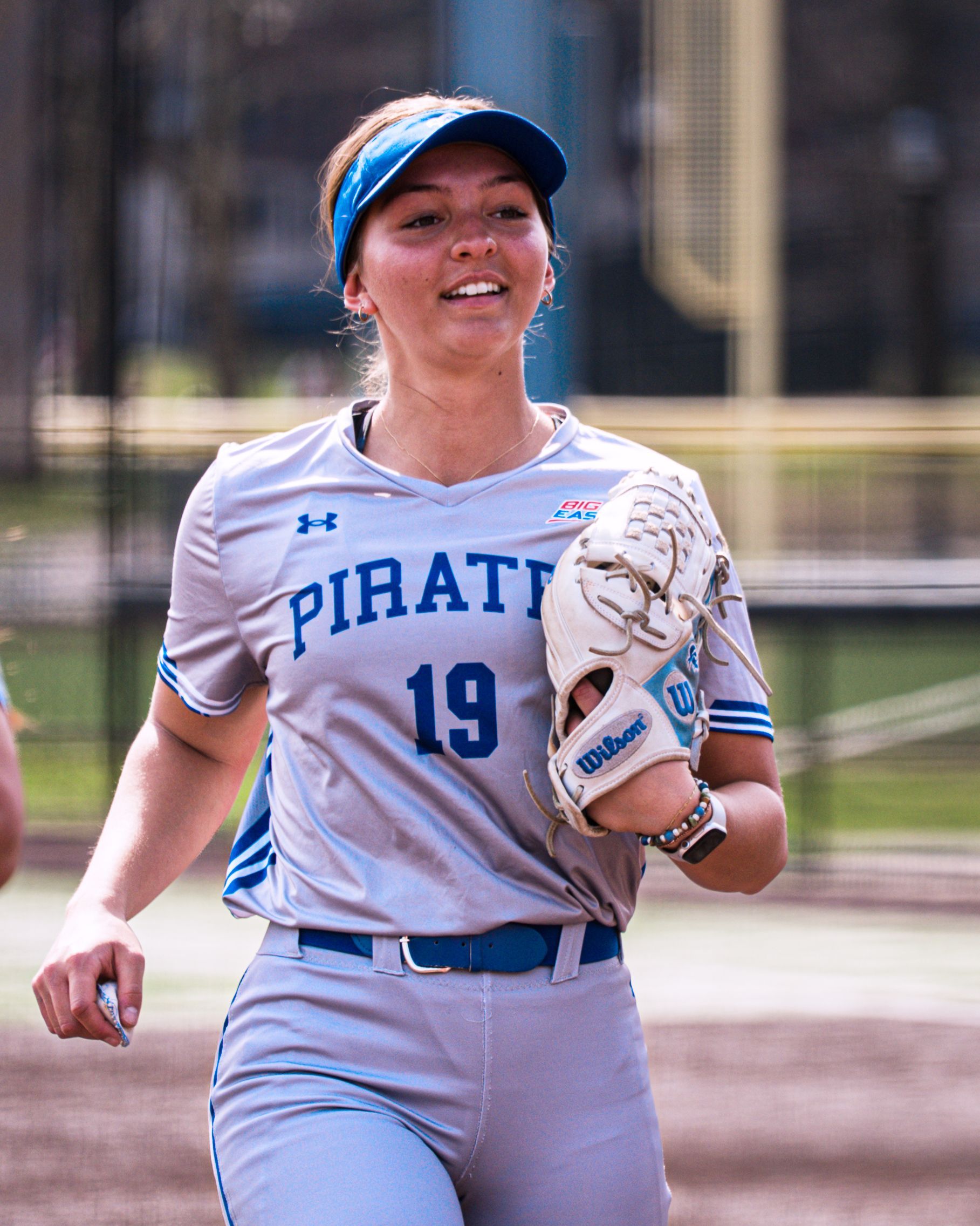 Softball player in a white Pirates uniform and blue visor walking on the field, holding a glove and smiling.