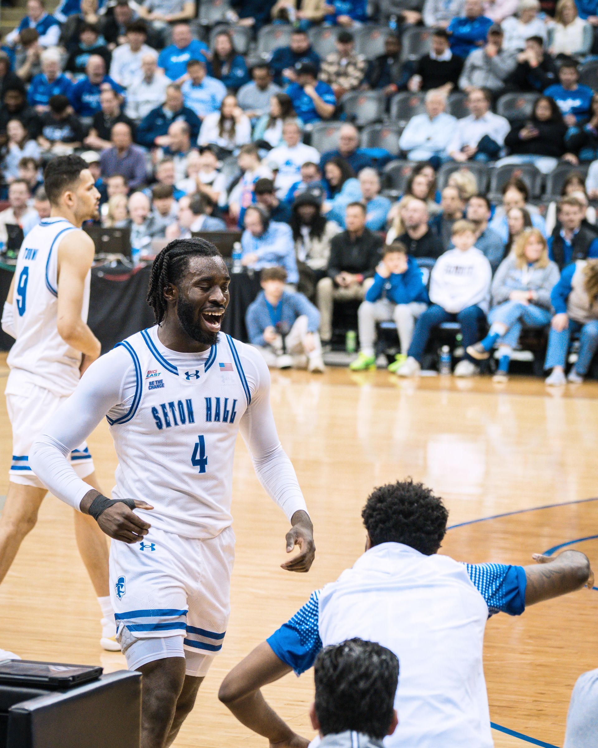 Basketball players in white and blue uniforms celebrate on the court with a cheering crowd behind them