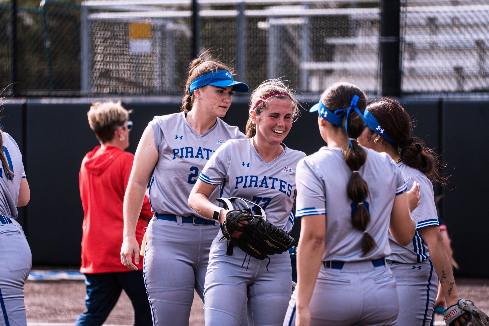 Softball players in gray uniforms huddle on the field, smiling and holding gloves.