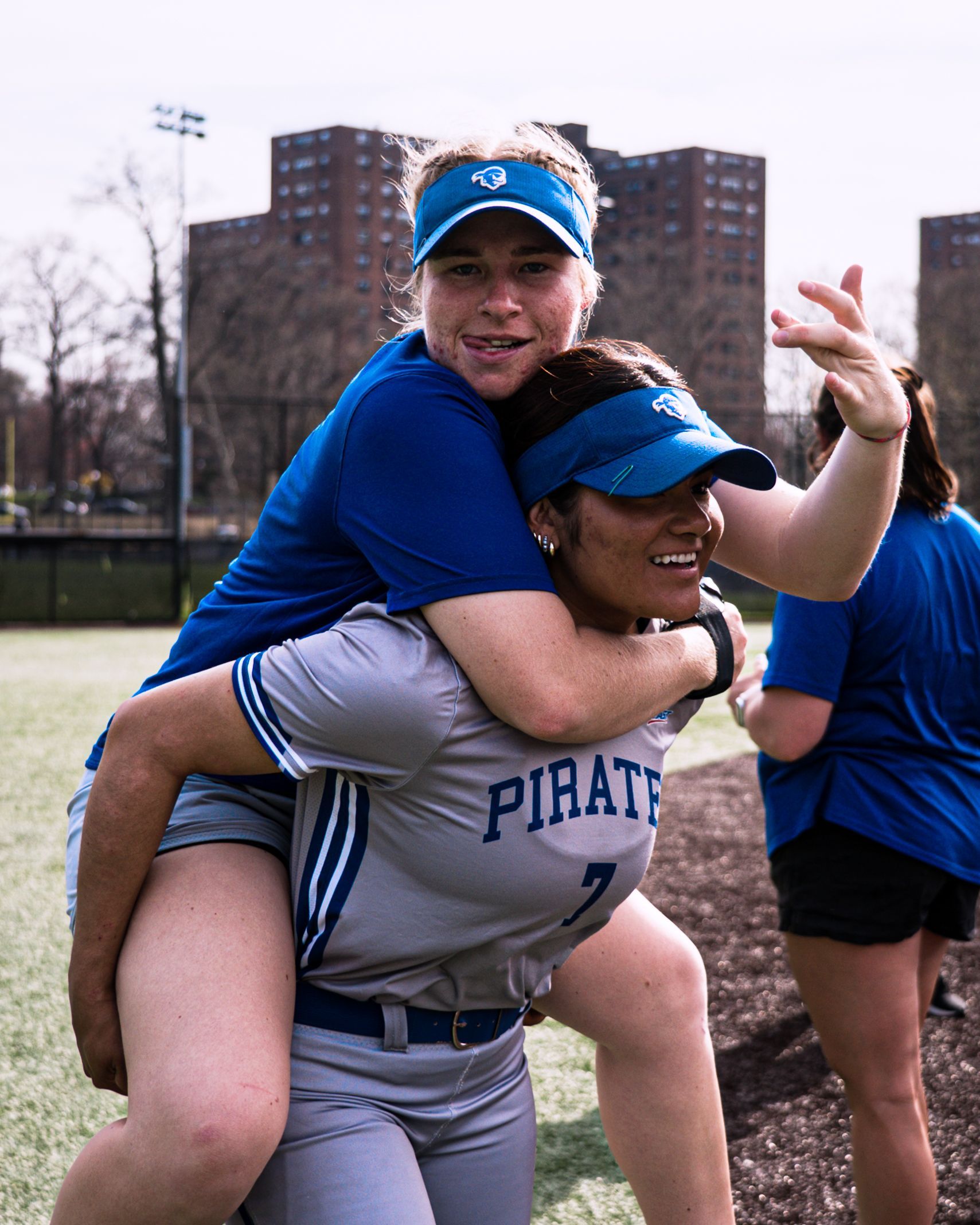 Two athletes in blue and gray uniforms, one giving the other a piggyback ride on a field.