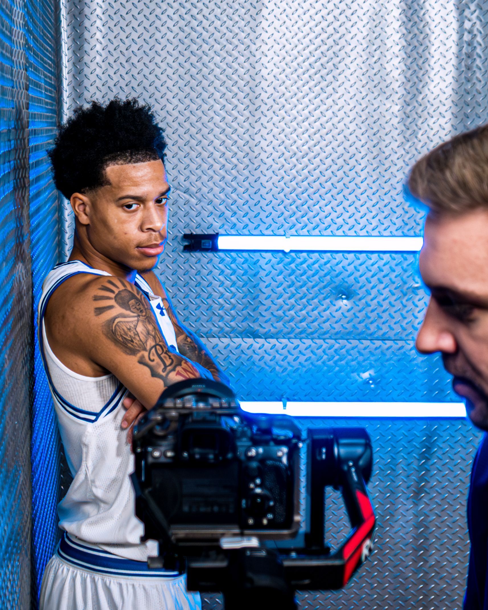 Man posing in a white jersey beside a camera, with blue neon lighting and a metallic backdrop.
