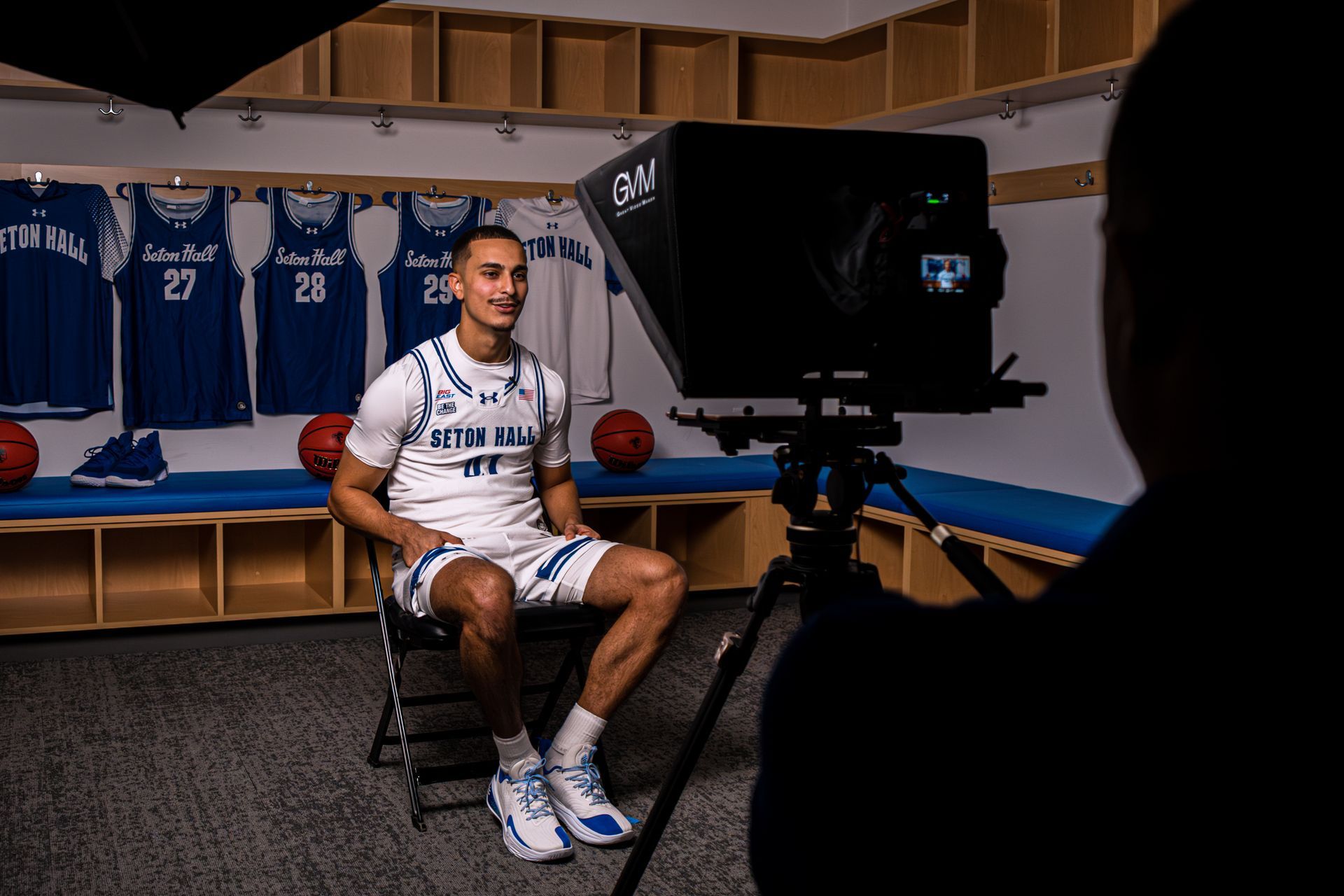 Athlete in white sports gear seated for an interview in a locker room, with blue jerseys behind him.