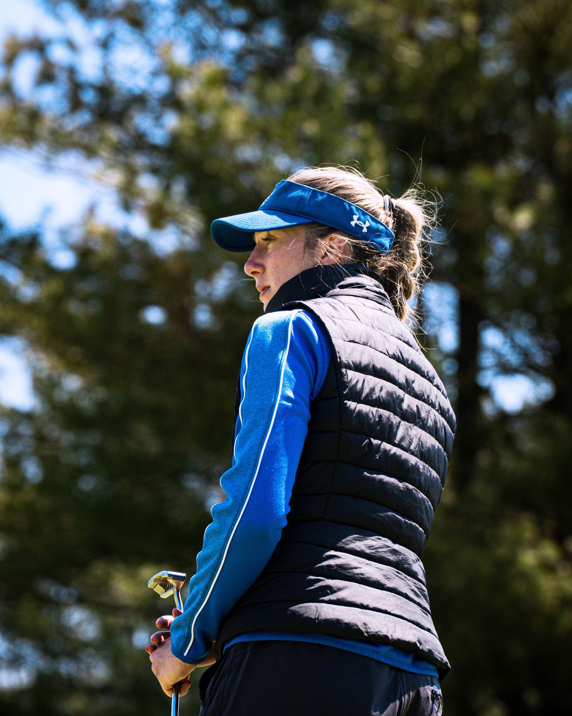Person in a blue visor and black vest standing outdoors near trees, holding a golf club