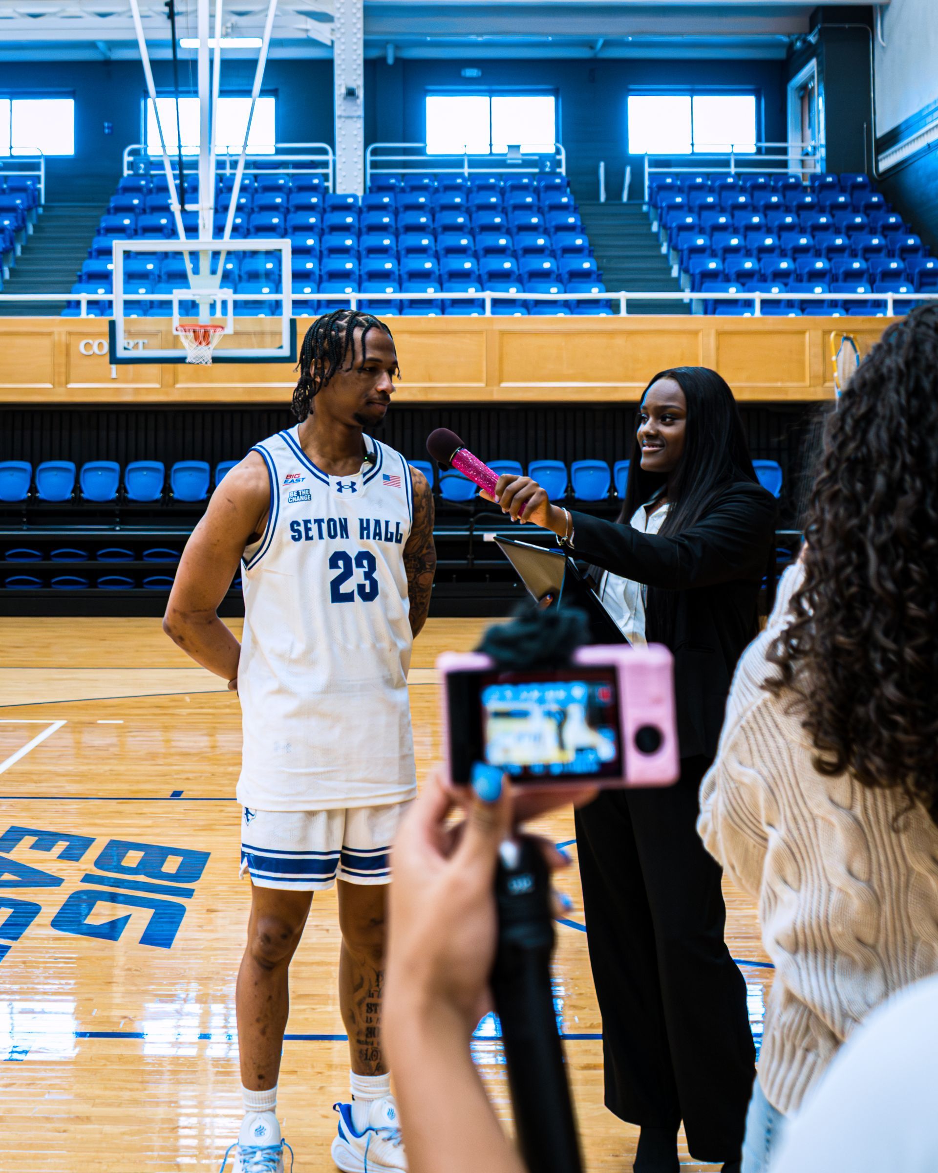 Basketball player in white jersey being interviewed on court with blue bleachers behind him