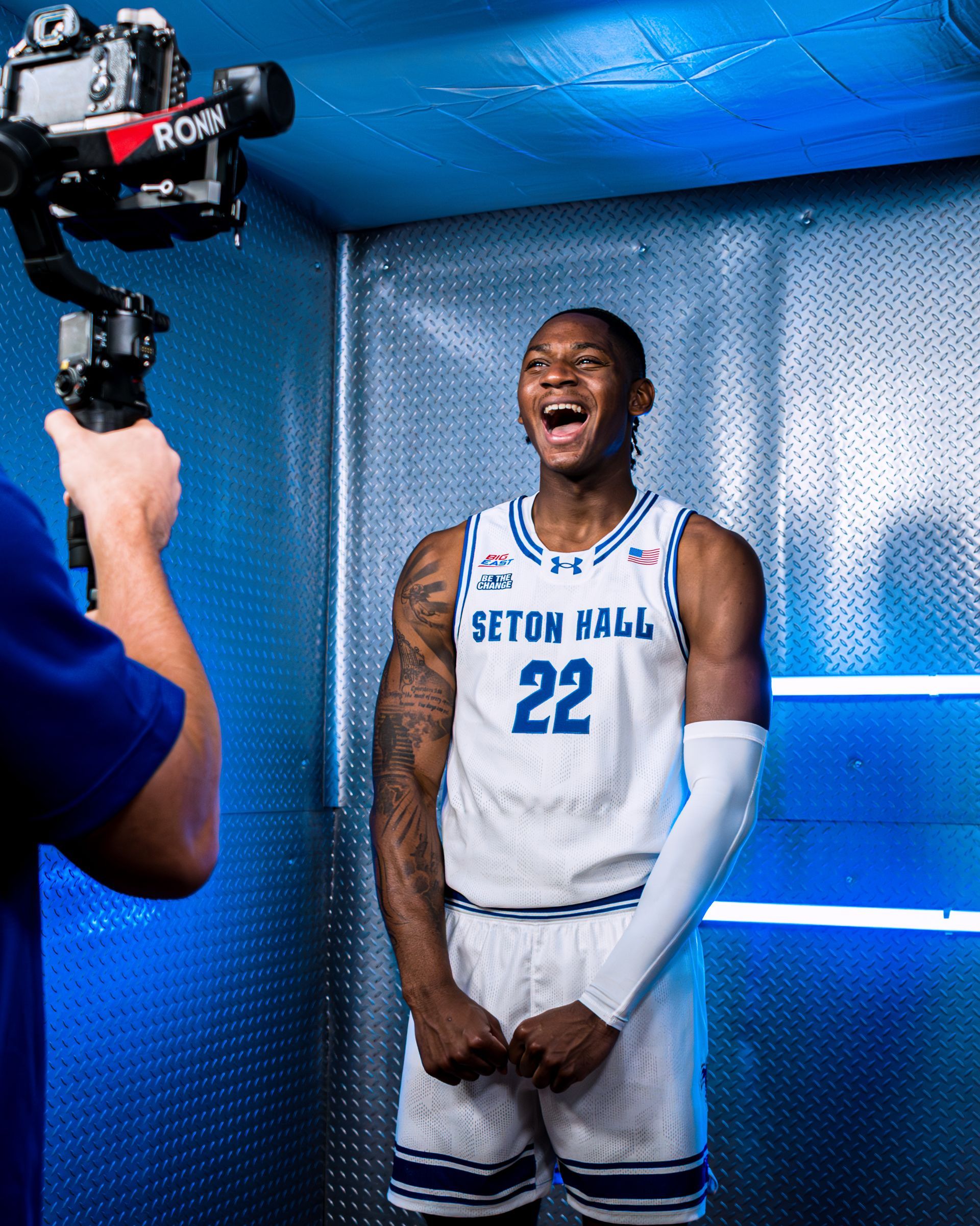 Seton Hall basketball player in white jersey posing and laughing in a blue-lit media backdrop
