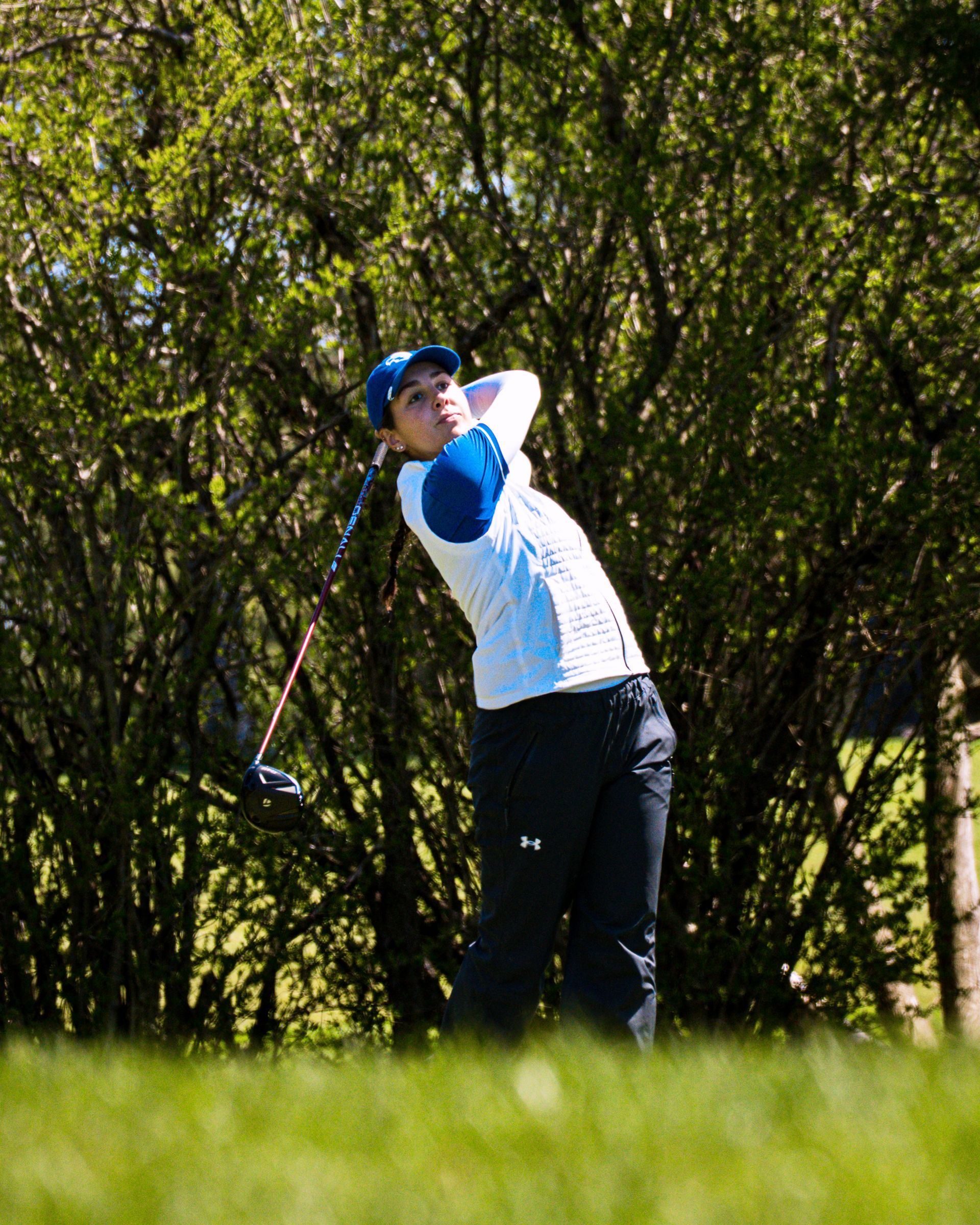 Golfer in blue and white swings on a green course with trees in the background