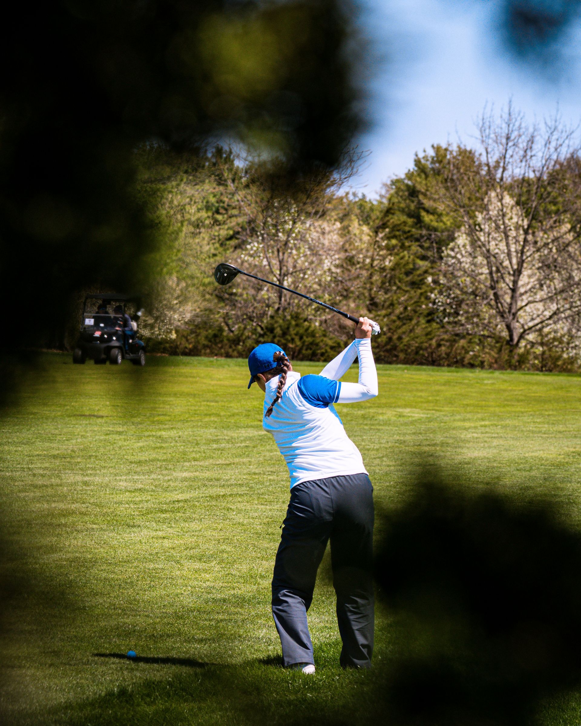 Golfer in blue cap and white shirt swinging a club on a green fairway with trees behind