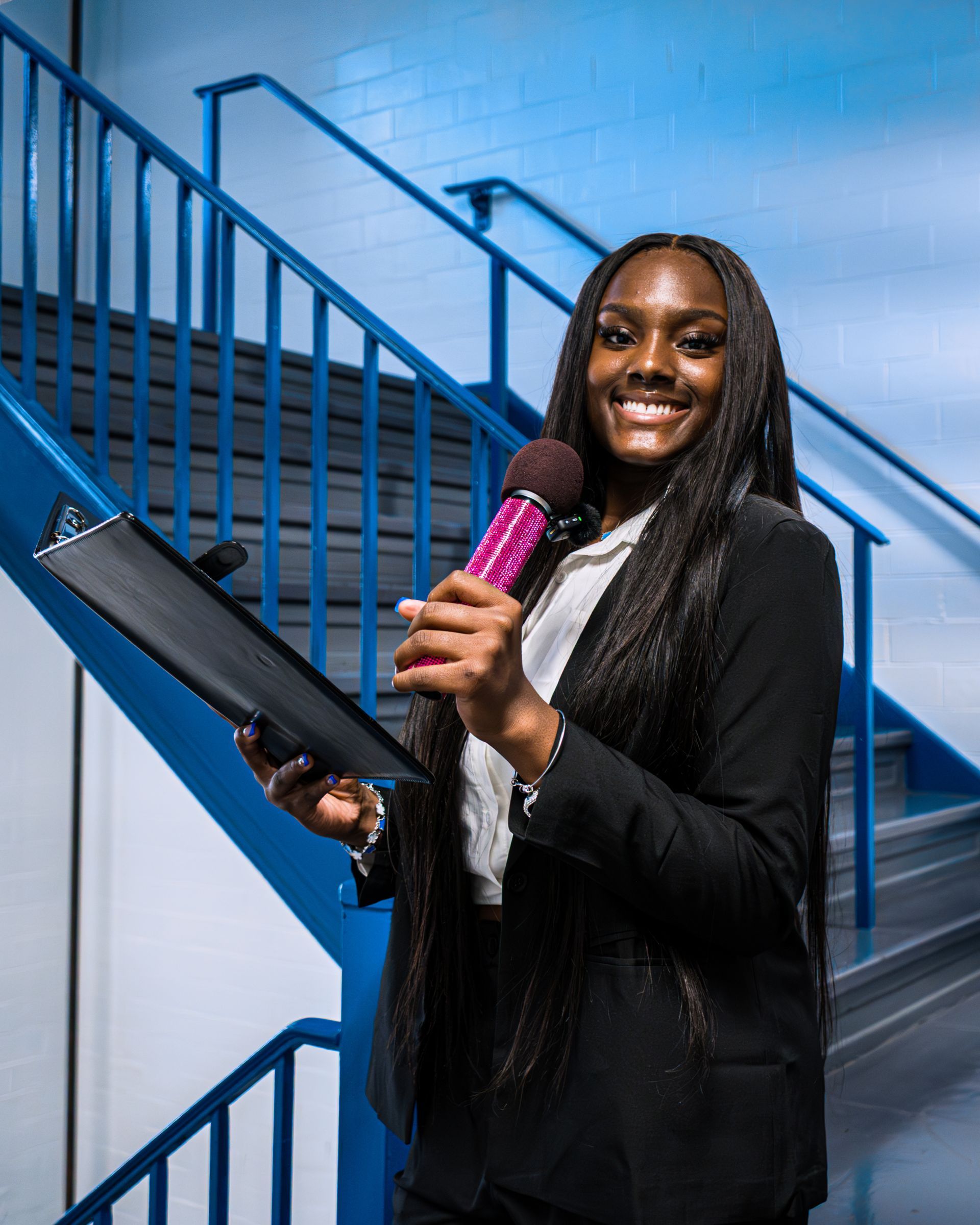Person smiling on a staircase holding a clipboard and pink microphone, wearing a black blazer.