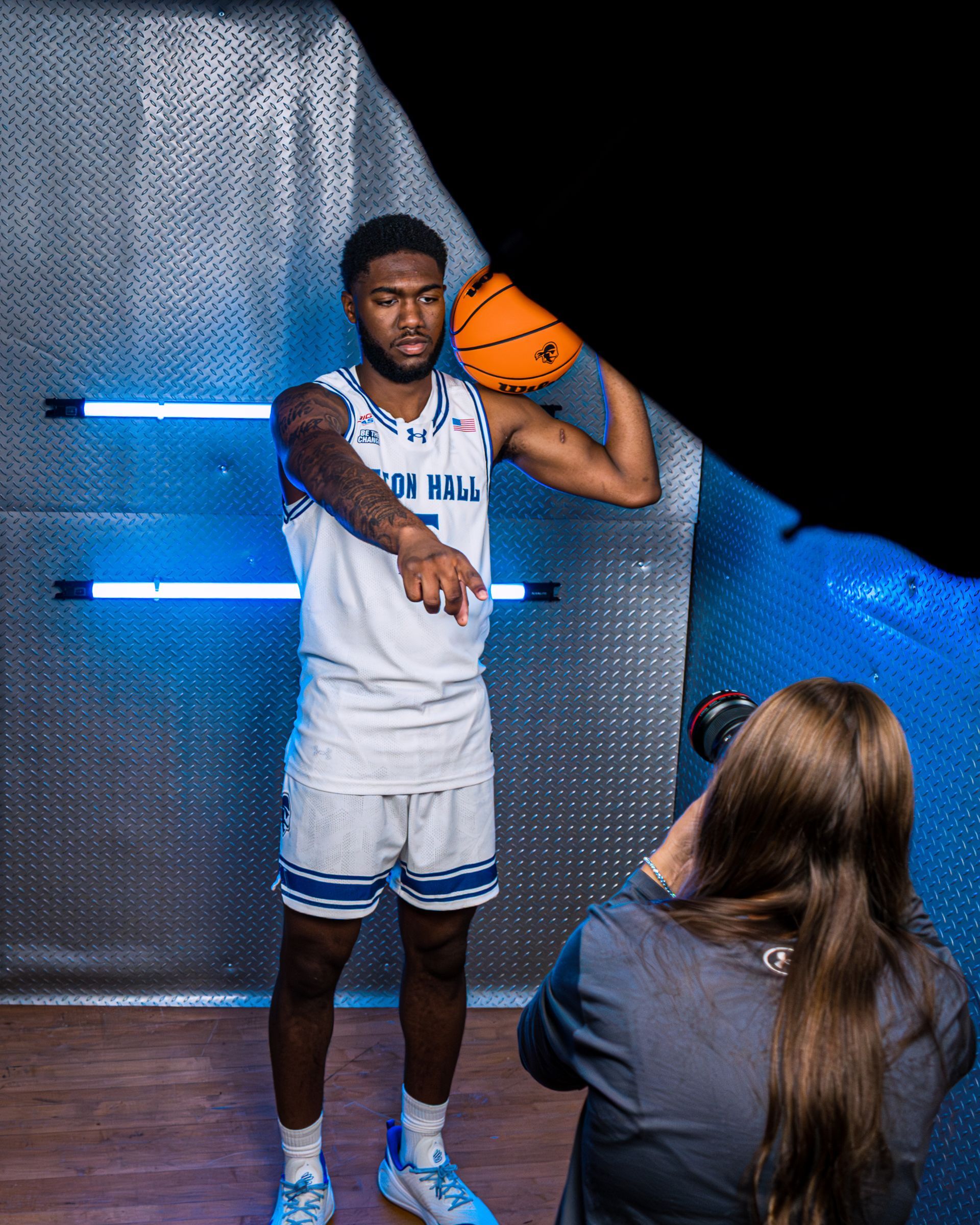 Basketball player in white uniform posing with a ball on a studio set while a photographer kneels nearby